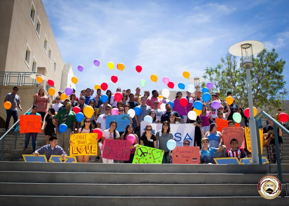 A group of people gathered on outdoor steps, holding colorful signs and lots of balloons gently blowing in the wind. 
