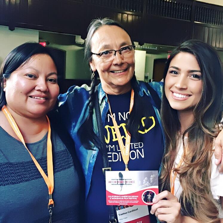 Missy Magooshboy, Tantoo Cardinal, Megan Doughty standing together with Megan Doughty holding a California's American Indian & Indigenous Film Festival informational card so that it can be seen by the camera