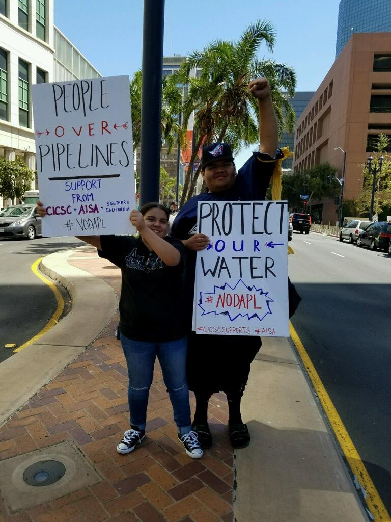 Two people holding signs that say, "People Over Pipelines, Support From CICSC + AISA + Southern California #DONAPL" and "Protect Our Water #DODAPL #CICSCSUPPORTS #AISA" 
