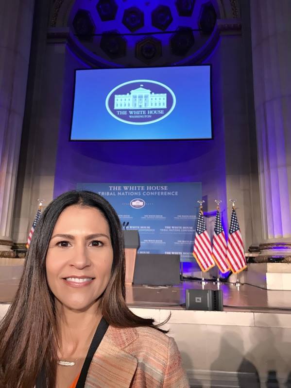 A selfie of Joely Proudfit standing in front of a stage with a large projector in the air that has the White House logo against a blue screen and below that is the stage, which has a podium in front of a large blue sign/background titled "White House Tribal Nations Conference" and there are American flags positioned on either side of the sign