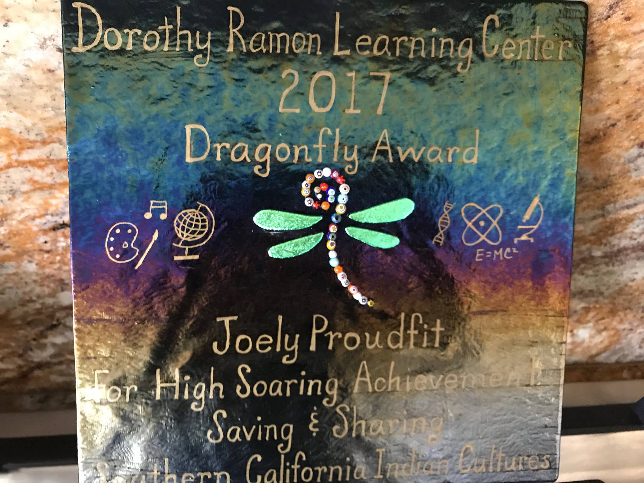 A plaque with the words, "Dorothy Ramon Learning Center 2017 Dragonfly Award Joely Proudfit For High Soaring Achievement Saving & Sharing Southern California Indian Cultures" and a large image of a dragonfly with sparkling green wings and a body made out of beads, and smaller symbols of music notes, a paint palette and brush, a globe, a DNA helix, an atom, a microscope, and the text, "E = MC^2" 
