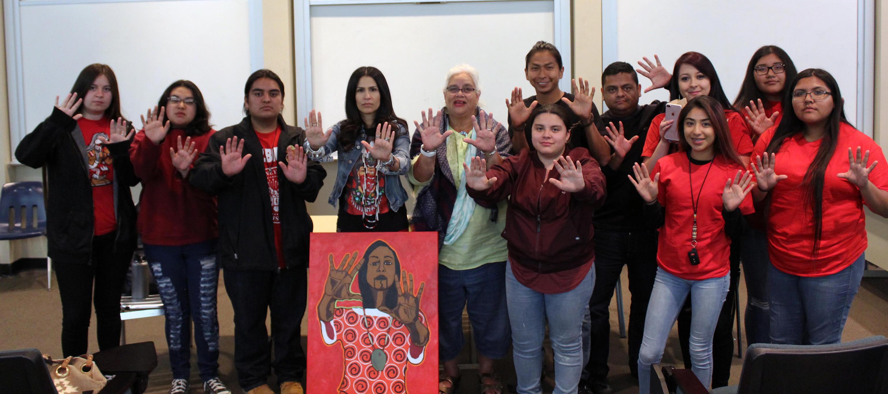People standing together behind a painting of a Native woman wearing red and holding both of her hands out, palms forward. The people who are standing have serious expressions and are also holding their hands out, palms forward, just like the woman in the painting.