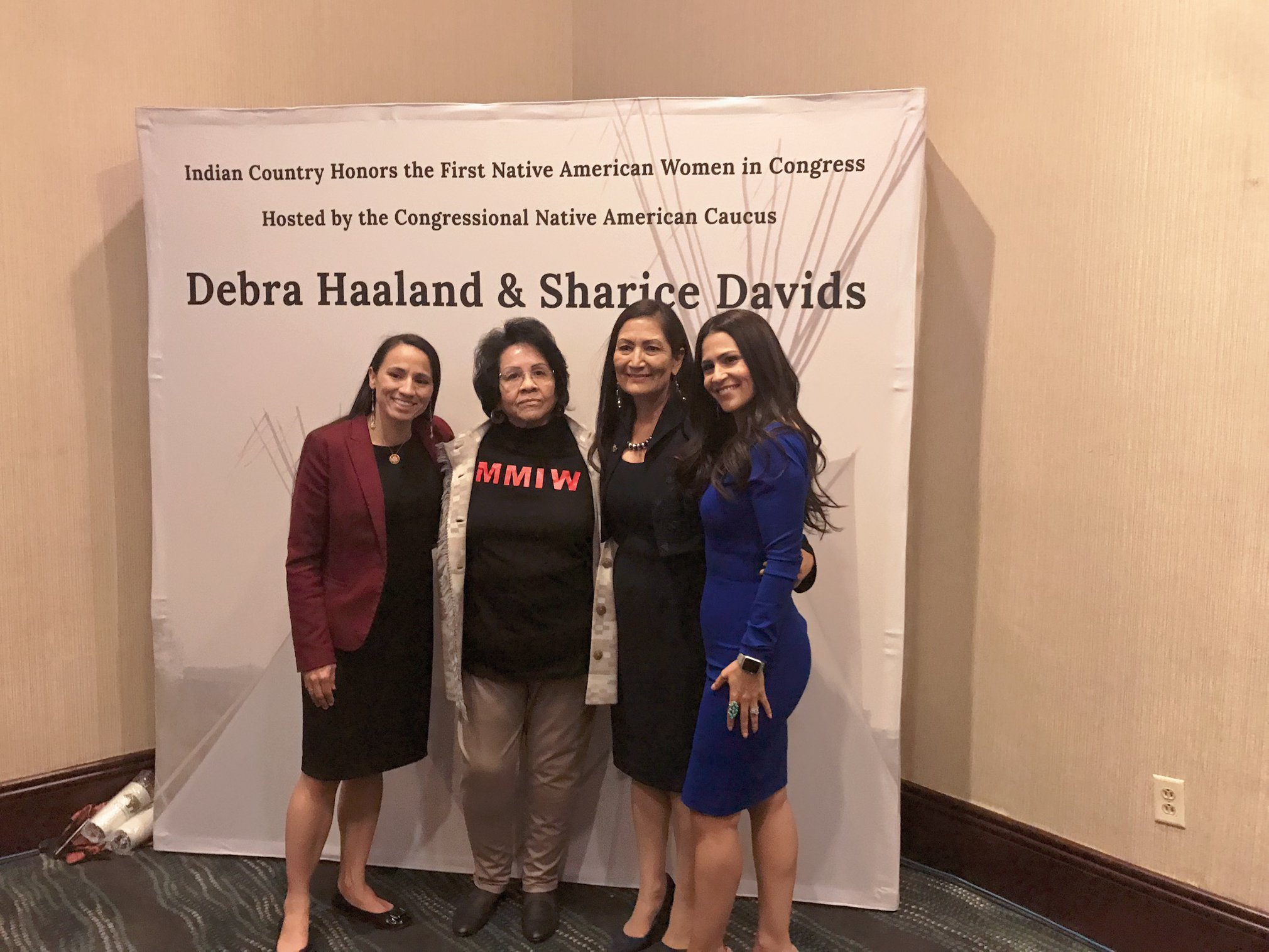 Sharice Davids, Mary Ann Martin Andreas, Debra Haaland, and Dr. Joely Proudfit standing together in front of a backdrop that states, "Indian Country Honors the First Native American Women in Congress Hosted by the Congressional Native American Caucus, Debra Haaland & Sharice Davids"