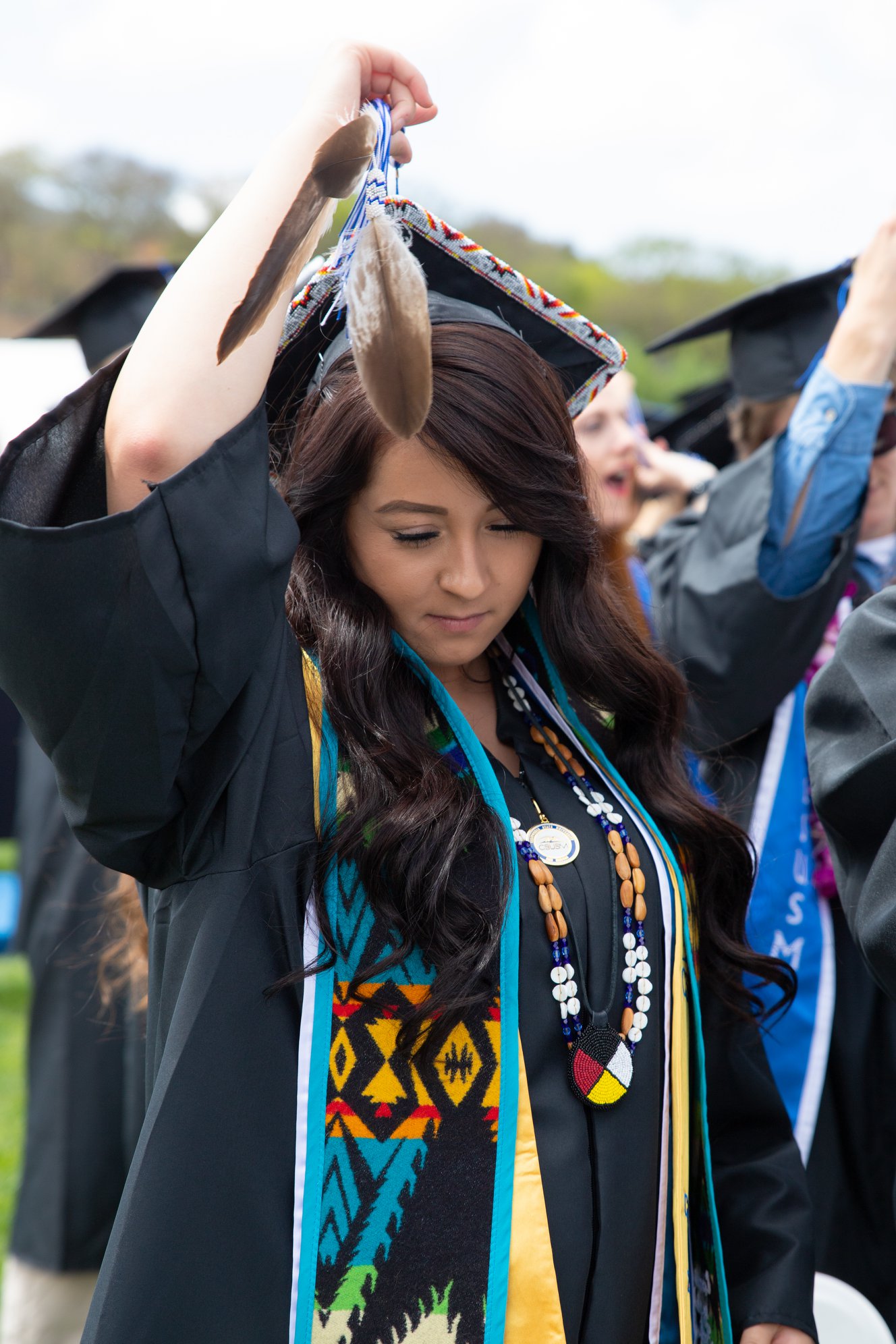 Graduate Rachelle Peterson in her cap and gown at graduation, wearing a multicolored pendleton stole and holding the feather on her graduation cap above her bowed head