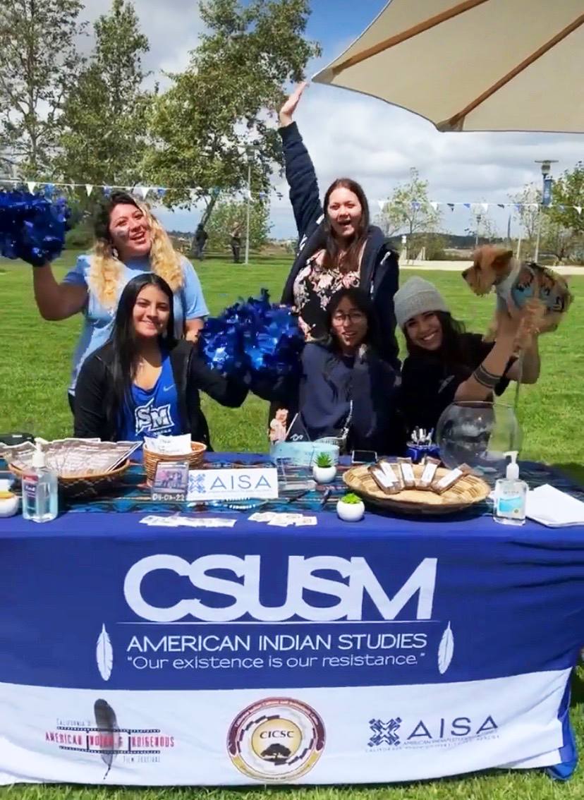 People sitting down at or standing behind a table outside on the grass. The table has a blue "CSUSM American Indian Studies 'Our existence is our resistance'" tablecloth that has a variety of materials on top. The people are cheering and smiling, two people are holding blue pom poms, and one person is holding up a little dog.