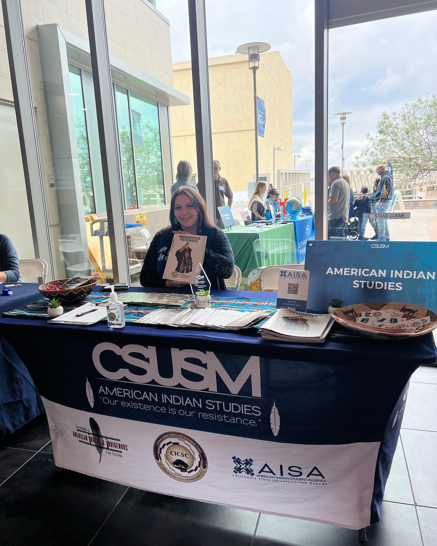 AI/AN student sitting at a CSUSM American Indian Studies table that contains pamphlets and other items, holding up an Indigenous Pathways packet