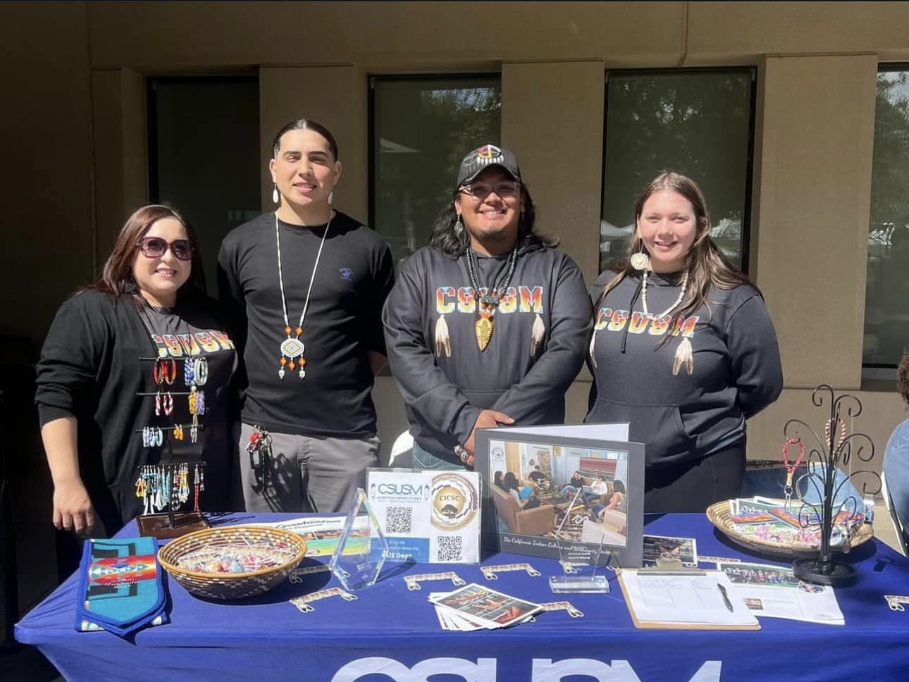 Four people standing outside behind a table with a blue tablecloth and a variety of items showcasing AI/AN presence on campus, including jewelry, a Pendleton stole, stickers, pamphlets, a sign-up sheet, and more