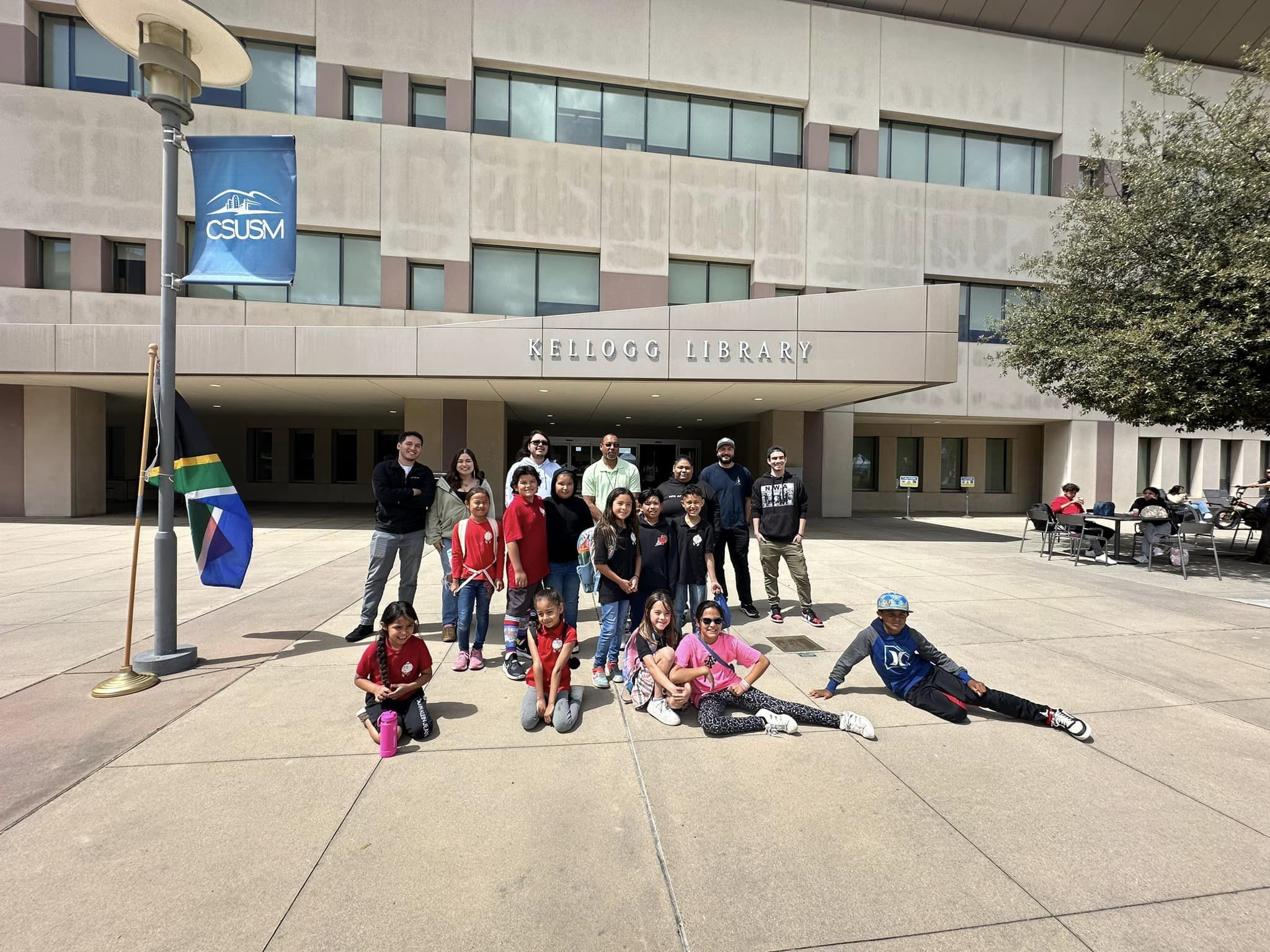 A group of children standing and sitting on the ground outside in front of the Kellog Library at CSUSM with staff and AISA students standing with them