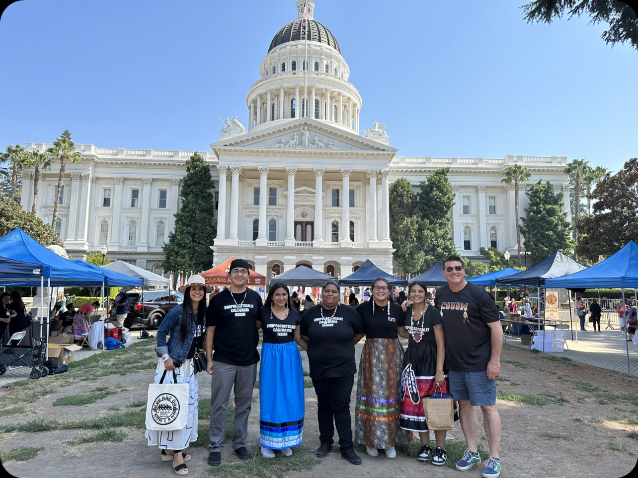 Students and faculty standing in a dirt courtyard that is surrounded by pop-up canopies and in front of the California State Capitol Building. The students and faculty are all wearing black shirts, some with the words, "Unapologetically California Indian," some with the letters, "CSUSM." 