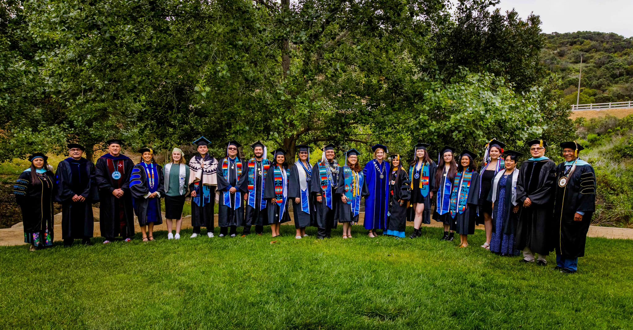 Graduates and faculty and staff in their caps, gowns, and stoles, standing in a horizontal line in the grass in front of a large green tree.