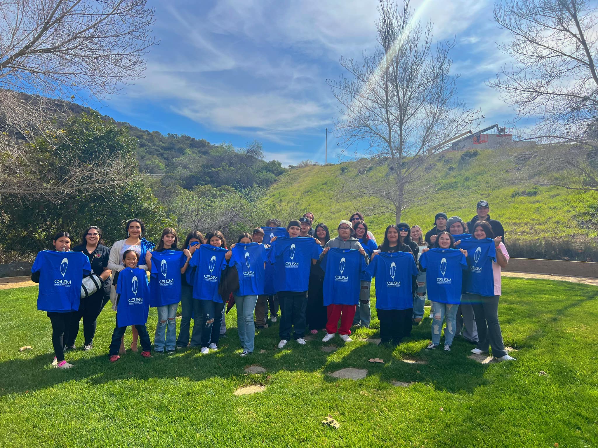 Group of children standing outside on bright green grass as they hold up blue CSUSM t-shirts that have a white feather graphic on them
