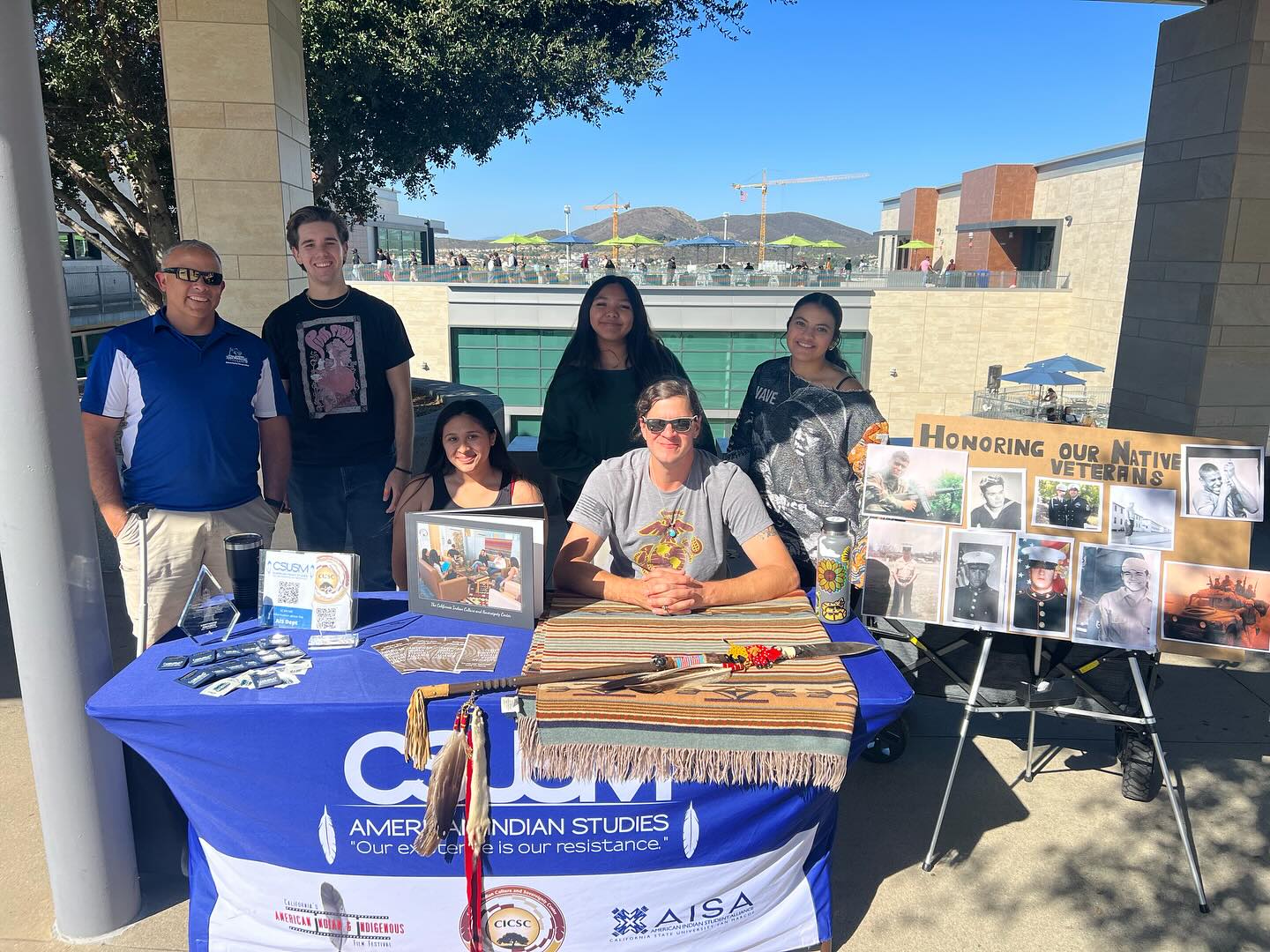 A group of people sitting at or standing behind a table with a blue American Indian Studies tablecloth on it as well as various items on the table and a sign on a stand to the side that has pictures of veterans and the words, "Honoring Our Native Veterans"