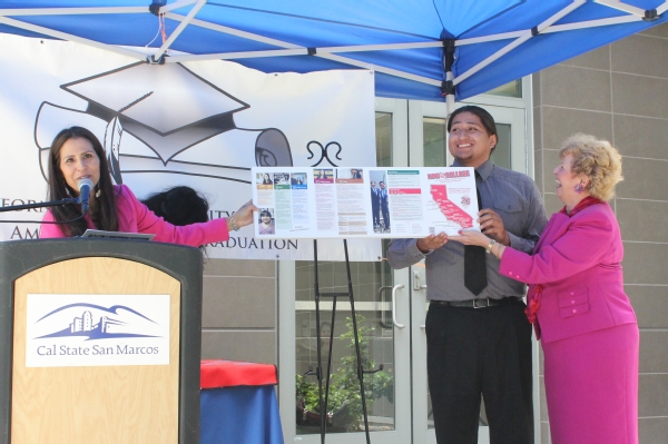 At an outdoor event beneath a blue pop-up tent, CICSC Director Dr. Joely Proudfit speaks into a microphone at a podium and holds up one side of the brochure while a Native American student holds up the other side with then-CSUSM president Karen Haynes 